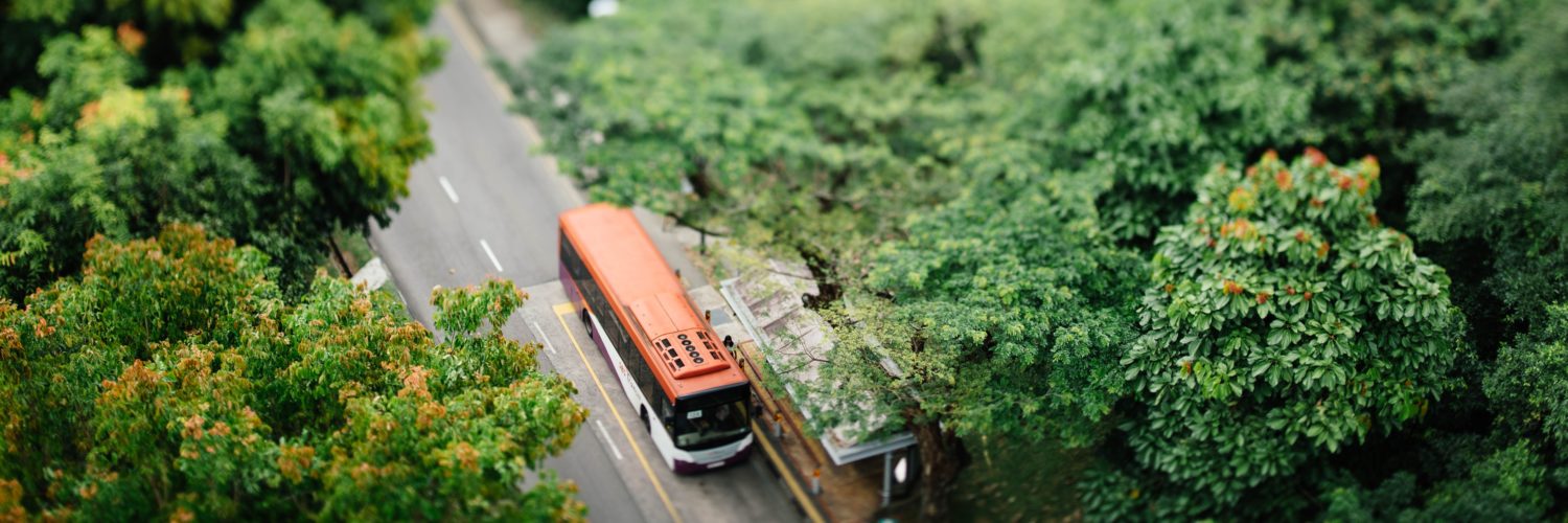 An aerial tilt-shift view of a bus stopped along a tree-lined street