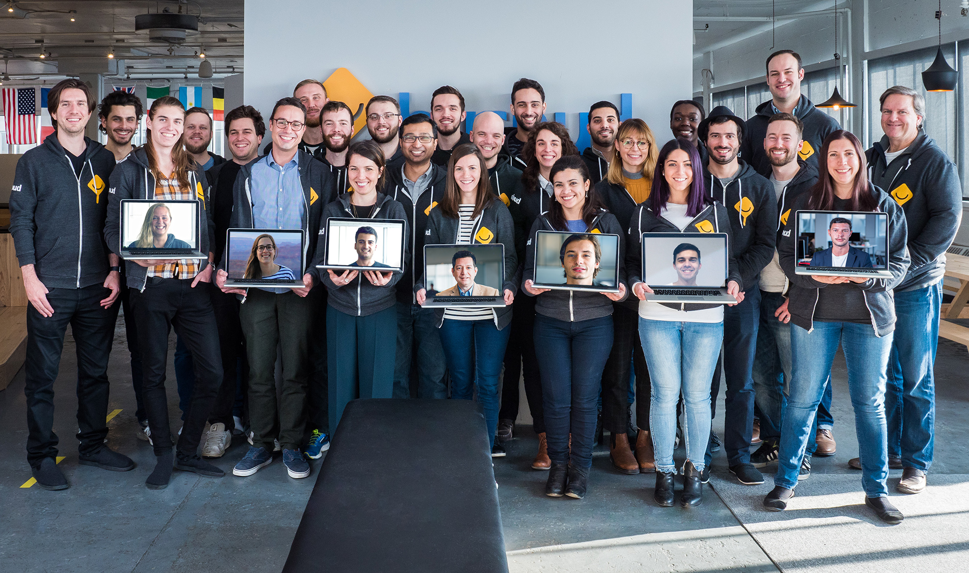The Busbud team posing together in the Montreal office, several members holding laptops displaying the faces of remote colleagues joining the photo