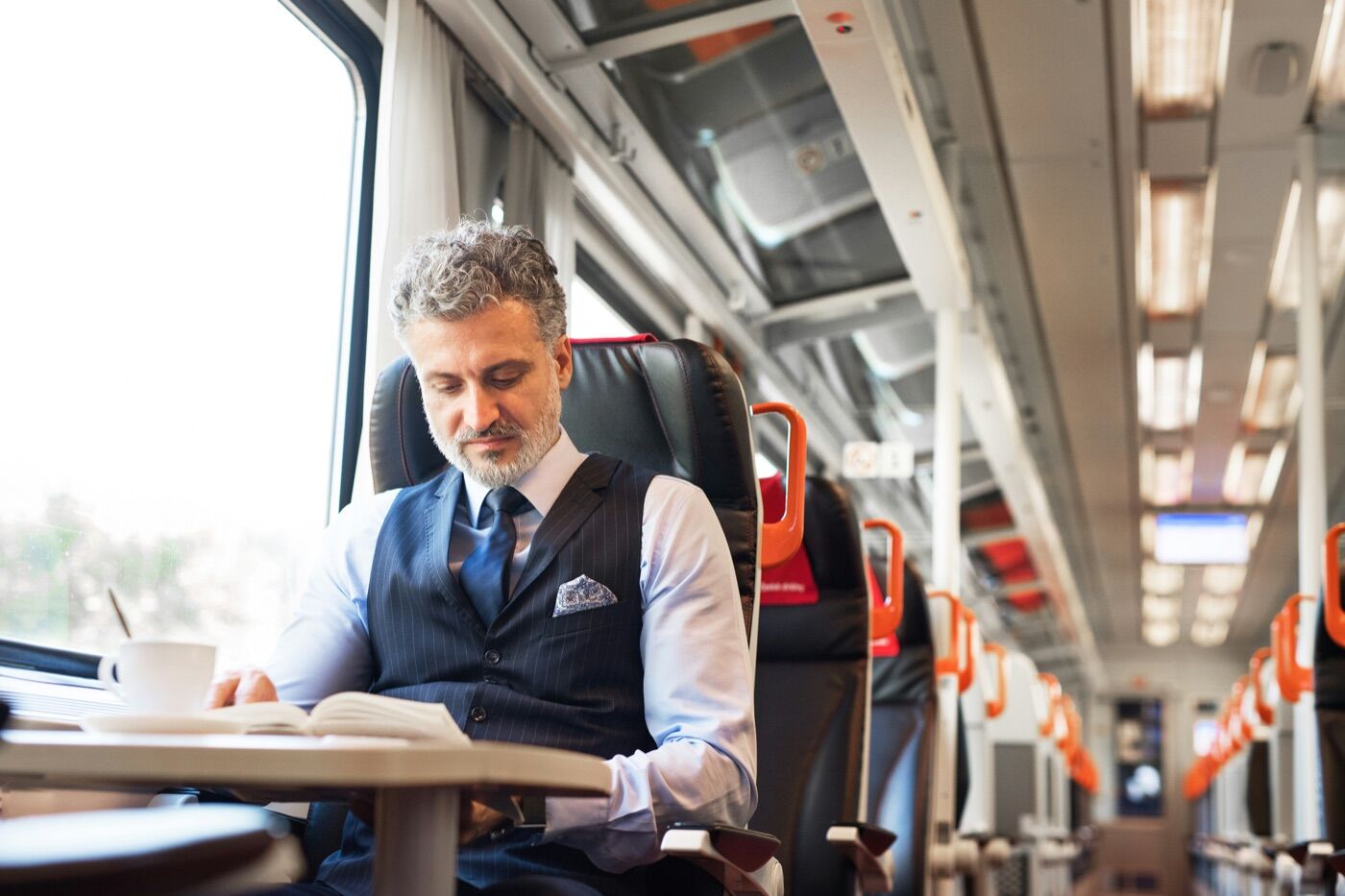 Well-dressed long-distance passenger reading at a train table with coffee, partner cabin extending behind him